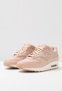 Tenisky Nike Air Max 1 z měkké růžové kůže a semiše, s texturovaným svrškem, polstrovaným límcem a viditelnou jednotkou Air v podešvi.