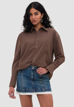 Vrouw met krullend zwart haar, zilveren oorringen, een bruin knoopjesoverhemd en een blauwe spijkerbroek minirok over een witte laag.