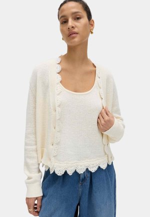 Donna che indossa un top a maglia color avorio con bordi in pizzo smerlato, abbinato a un cardigan con bottoni dello stesso colore e jeans blu larghi.