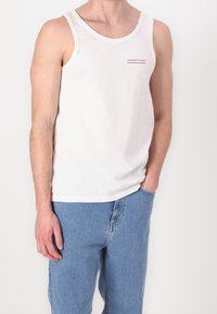 Hombre con camiseta sin mangas blanca de Tommy Jeans y jeans azul claro, mano en el bolsillo, contra un fondo blanco liso.