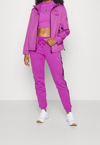 Kvinna iklädd en ljuslila crop-top, matchande joggingbyxor och en lätt jacka med vita sneakers, som står mot en vit bakgrund.