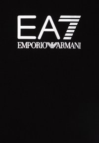 Melnas audums ar atstarojamām baltām vēstulēm, kurās ir ierakstīts "EA7 EMPORIO ARMANI", un dizaina horizontālās svītras numura 7 formā.