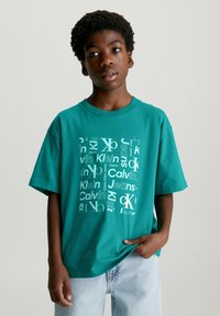 Camiseta de algodón color azul verdoso con un estampado gráfico que presenta varios logotipos de Calvin Klein en blanco. Mangas cortas y corte relajado, combinada con jeans de mezclilla claros.