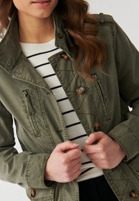 Chaqueta verde oliva con un cuello alto, cuatro bolsillos y cierres de botón. El modelo lleva una camiseta a rayas debajo. Tejido texturizado.