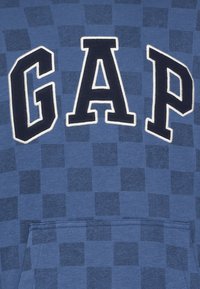 Felpa blu con motivo a scacchi. Grande logo "GAP" in blu navy, contornato in bianco. Presenta una tasca frontale e una texture di tessuto morbido.