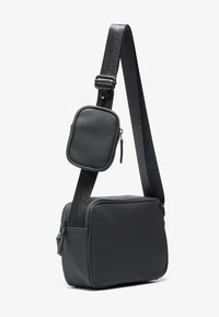 Borsa a tracolla nera realizzata in materiale liscio, con una mini tasca staccabile, tracolla regolabile e forma rettangolare. Design minimalista.