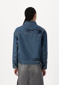 Chaqueta de denim en azul claro, con bordados florales en la parte posterior y en las mangas. Cuello clásico y cierre de botones, con una silueta ajustada.