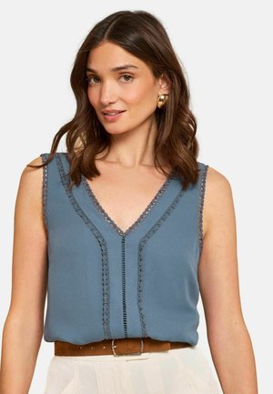 Vállig érő barna hajú nő arany karika fülbevalóval, ujjatlan kék felső díszítő szegéllyel és barna övvel fehér nadrágon.