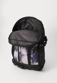 Mochila negra con un exterior texturizado, que cuenta con un gran bolsillo frontal en patrones abstractos de azul y rosa, y una solapa superior abierta con cremallera.