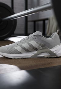 Chaussure de sport grise avec des rayures blanches reposant sur le sol de la salle de sport, avec une barre et des poids en arrière-plan flou.