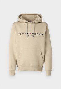 Beige hoodie in soft fabric with a front pocket. Features "TOMMY HILFIGER EST. 1985" logo in navy, and white drawstrings at the hood.