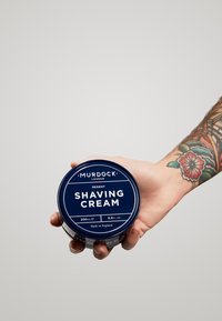 Barattolo di metallo blu di crema da barba Murdock Regent, etichettato in modo evidente, tenuto in una mano, con una superficie liscia e tipografia nera.