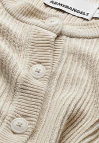 Cardigan en tricot côtelé couleur crème avec des boutons blancs embossés "ARMEDANGELS" et une étiquette de marque visible à l'intérieur du col.