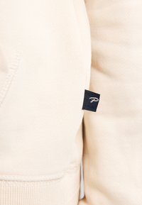 Felpa beige chiaro con una texture morbida, polsini a costine e un'etichetta con logo ricamato nera sul lato.