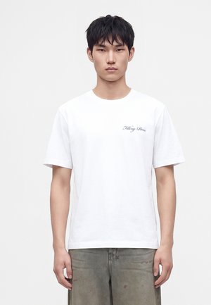 Jeune homme aux cheveux courts noirs portant un T-shirt blanc avec un petit texte noir sur la poitrine gauche et un pantalon gris avec des taches.