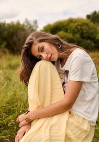Jeune femme en t-shirt graphique blanc et jupe jaune assise sur l’herbe, reposant sa tête sur son genou, avec des arbres en arrière-plan.