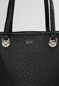 Sac à main noir Guess avec motif embossé, double bandoulière et détails en métal argenté avec texte du logo « GUESS EST 1981 ».