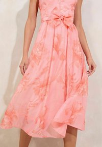 Pink floral dress featuring a fitted bodice, bow detail at the waist, and a flowing skirt with a light, sheer texture and subtle pattern.