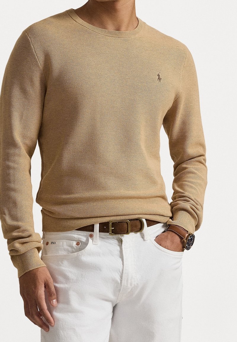 Homme portant un pull texturé beige à manches longues avec un petit logo, pantalon blanc, ceinture en cuir marron et montre, debout avec une main dans la poche.