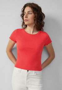Vrouw met krullend haar, gekleed in een strak, rode top met korte mouwen en witte broek, staand met de handen achter de rug tegen een grijze muur.