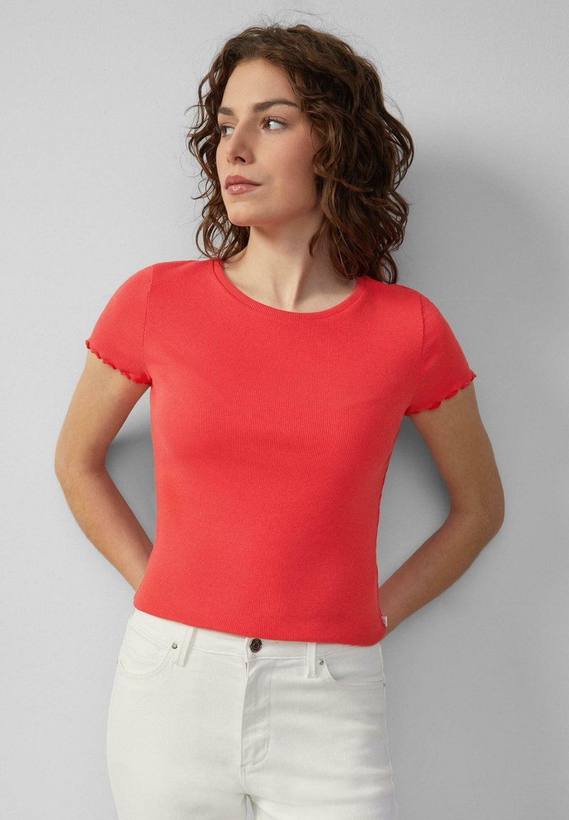 Vrouw met krullend haar, gekleed in een strak, rode top met korte mouwen en witte broek, staand met de handen achter de rug tegen een grijze muur.