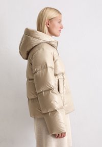 Beżowa pikowana kurtka z kapturem, o błyszczącej fakturze, miękkim materiale, pudełkowym kroju i bocznych kieszeniach, widoczna z prawej strony.