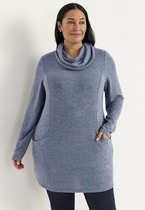 Kvinde iført en blå sweater med lange ærmer og løsthængende krave med lommer, stående mod en ensfarvet lys baggrund.