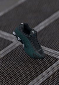 Sapatilhas Nike em preto e verde escuro com uma superfície texturizada. As características incluem um cabedal em malha, cordões proeminentes e um acolchoamento distinto no calcanhar.