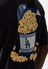 T-shirt noir arborant un grand graphique d'un seau de pop-corn avec des rayures bleues et blanches, surmonté de pop-corn coloré et d'un logo LA.