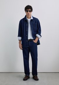 Man in een donkerblauwe denim jas en broek over een gestreept shirt en witte t-shirt, met zwarte zonnebril en bruine schoenen, staande binnen.