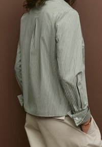 Striped long-sleeve shirt in olive green and white, featuring a back pleat, button-down cuffs, and a relaxed silhouette. Light fabric texture.