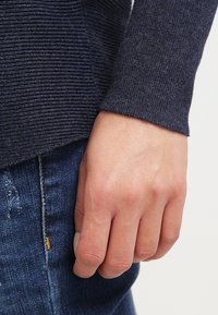 Maglione a maniche lunghe a coste color blu navy con una finitura tessurizzata, abbinato a un paio di jeans in denim blu. In primo piano, una mano appoggiata sulla tasca dei jeans.