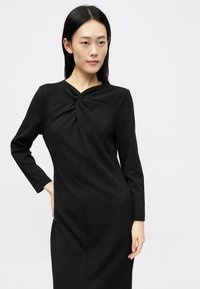 Crna haljina dugih rukava sa dizajnom uvijenog čvora na dekolteu. Glatka tkanina s uskim siluetom, bez šavova i minimalistička.