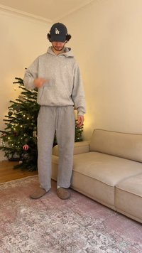 Szara bluza z kapturem i kieszenią kangurka, dopasowane szare spodnie dresowe oraz beżowe kapcie. Stoi w pokoju z choinką.