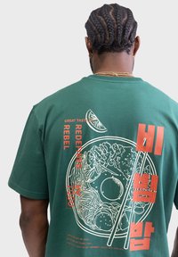 Grünes Baumwoll-T-Shirt mit einer weißen illustrierten Schüssel mit Nudeln und Essstäbchen, akzentuiert durch roten Text und eine Limette-Grafik.