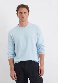 Jasnoniebieski sweter crewneck z długimi rękawami, wykonany z miękkiego materiału. Noszony z ciemnymi spodniami, o luźnym kroju i ściągaczach przy mankietach.