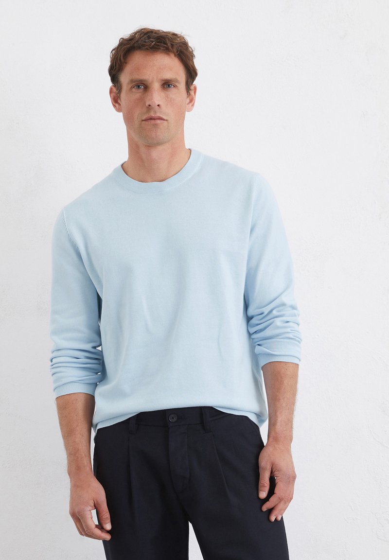 Jasnoniebieski sweter crewneck z długimi rękawami, wykonany z miękkiego materiału. Noszony z ciemnymi spodniami, o luźnym kroju i ściągaczach przy mankietach.