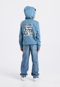 Sweatshirt à capuche bleu avec des noms de villes urbaines imprimés en noir et blanc dans le dos. Associé à un jean baggy bleu clair et des baskets blanches.