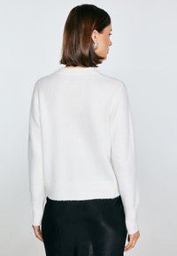 Bílý pletený svetr s texturou, dlouhými rukávy a zkráceným střihem. Má žebrované manžety a lem, nošený přes hladký černý oděv.