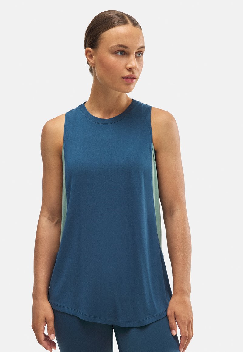 Mujer con el cabello recogido, vistiendo una camiseta sin mangas azul y leggings a juego, mirando hacia su derecha frente a un fondo blanco liso.