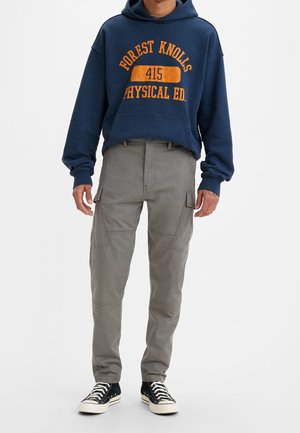 Personne portant un sweat à capuche bleu marine « Forest Knolls Physical Ed », un pantalon cargo gris et des baskets noires et blanches, debout sur fond blanc.