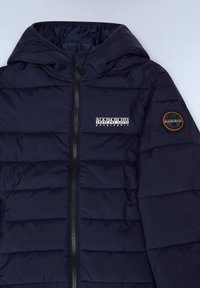 Mörkblå vadderad jacka med hög krage, full dragkedja och synliga quiltade sektioner. Har en logotyp på vänster ärm.