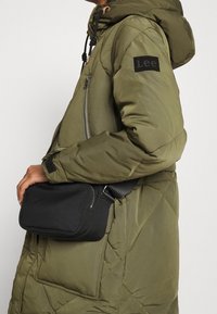 Chaqueta de plumón color verde oliva con textura acolchada y parche de logo negro en la manga, combinada con un pequeño bolso negro de bandolera.