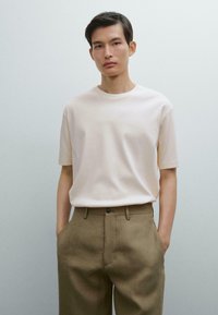 T-shirt a maniche corte color beige chiaro con scollo rotondo, indossato con pantaloni a vita alta verde oliva dotati di tasche laterali e chiusura con bottone.