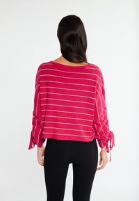 Fuktsia neulepaita, jossa on vaakasuoria hopeanvärisiä raitoja, ylisuurta mitoitusta ja kootut hihat, jotka on sidottu ranteisiin. Pehmeä neulekangas.