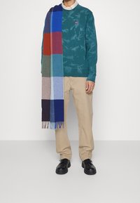 Teal sweatshirt met Levi's logo, crème kraag, beige broek en een multicolore geruite sjaal met franjes. Zwarte veterschoenen.