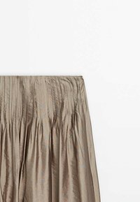 Jupe plissée beige brillante avec des détails froncés à la taille, suspendue devant un fond blanc.