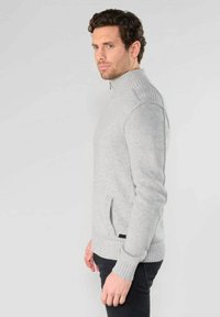 Grauer Strickpullover mit hohem Kragen und gerippten Bündchen. Verfügt über eine seitliche Reißverschlusstasche und eine glatte Textur, taillierte Passform.