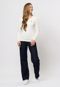 Bluză albă cu mânecă lungă și textură cord, accent floral mic, asortată cu blugi negri cu croială largă și adidași albi, pe un fundal neutru.