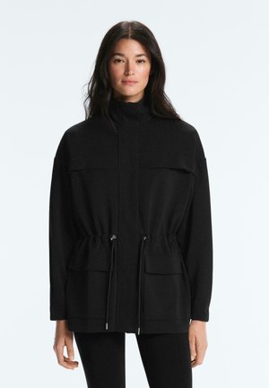 Nő fekete cipzáras dzsekit visel, melyen húzózsinóros derékrész és patentos zsebek találhatók, egyszínű világos háttér előtt áll.